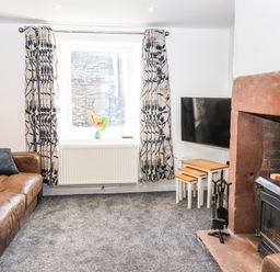 A living room with a brown leather sofa a window with patterned curtains a fireplace with a wood stove a flat screen tv and nesting tables at Sparrow Cottage in Keswick