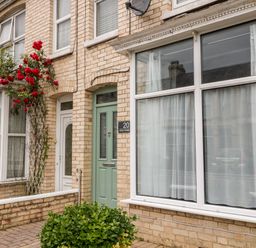 The exterior of a brick house with a green front door red flowers on a trellis and white framed windows with curtains at Daisy Cottage in Bideford
