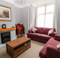 A living room with two red sofas floral cushions a wooden coffee table a fireplace and a wooden cabinet at Daisy Cottage in Bideford