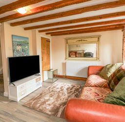 A living room with a patterned sofa and cushions a wall mirror a television on a stand and wooden ceiling beams at The Linhay in Holsworthy