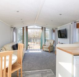 A living room with a sofa, armchair, dining table, wall-mounted television, and glass doors opening to a balcony at 109 Pentney Lakes Pentney near King's Lynn
