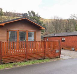 A wooden lodge with a fenced porch and French doors next to a similar lodge in a rural area at Lakeland View Lodge Beckside 2 Limefitt Park near Windermere