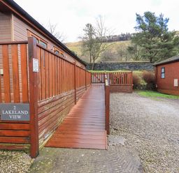 A wooden deck ramp leading to a cabin with a gravel driveway and trees in the background at Lakeland View Lodge in Beckside 2 Limefitt Park near Windermere
