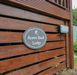 A wooden fence with a sign reading Acorn Bank Lodge near greenery and a building at Acorn Bank Lodge L 55 Fallbarrow Park near Bowness-On-Windermere