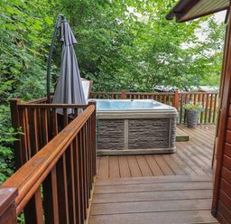 A wooden deck with a hot tub an umbrella and a potted plant surrounded by trees at Acorn Bank Lodge L 55 Fallbarrow Park near Bowness-On-Windermere