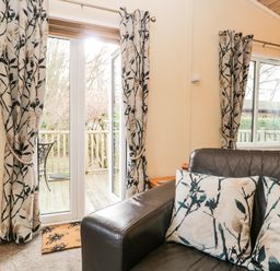 A living room with a leather sofa and floral patterned curtains by glass doors and a window at Damson Lodge Thirlmere 8 White Cross Bay Windermere