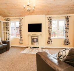 A living room with two brown leather sofas a wall-mounted television and fireplace two windows with patterned curtains and a wooden bookshelf at Damson Lodge Thirlmere 8 White Cross Bay Windermere