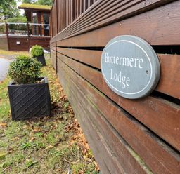 A wooden building with a sign reading Buttermere Lodge and potted bushes outside at Buttermere Lodge in Glade 5 Fallbarrow Park near Bowness on Windermere