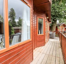 A wooden deck with railings outside cabins with large windows and a small table with chairs at Buttermere Lodge in Glade 5 Fallbarrow Park near Bowness on Windermere