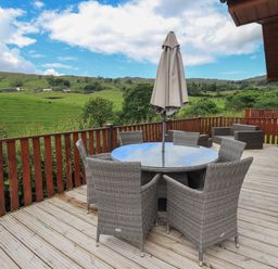 An outdoor wooden deck with a round glass table and six wicker chairs with a closed umbrella and two wicker armchairs overlooking green hills at Wansfell Retreat Lodge Wansfell 4 Limefitt Park near Windermere