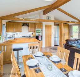 A dining area with a set table and an open kitchen with wooden cabinets and beams with a living area with leather sofas at Sheffield Pike Lodge Ullswater 8 Limefitt Park near Windermere