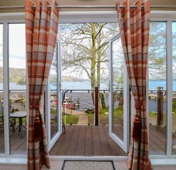 A room with open glass doors leading to a wooden deck overlooking a lake with trees and docks at Shoreside Lodge in L8 Fallbarrow Park near Bowness on Windermere