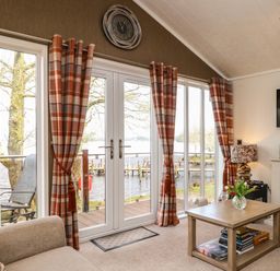 A living room with sliding glass doors and orange plaid curtains next to a wooden table with a vase of flowers at Shoreside Lodge in L8 Fallbarrow Park near Bowness on Windermere