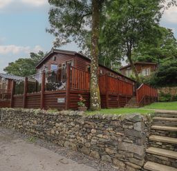 A wooden lodge with glass and wood railing on a raised stone platform with stairs and trees in the background at Shoreside Lodge L8 Fallbarrow Park near Bowness on Windermere