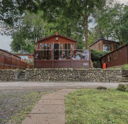 A wooden lodge with glass doors and a stone wall in front surrounded by trees at Shoreside Lodge L8 Fallbarrow Park near Bowness on Windermere