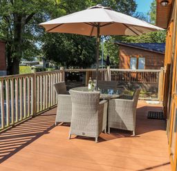 An outdoor deck with a round glass table seating four wicker chairs and a large umbrella at Daffodils Lodge in Oakside 7 Fallbarrow Park in Bowness on Windermere