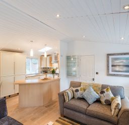 A living room with a sofa and cushions next to a kitchen area with a wooden breakfast bar at Broad Larch Lodge in Glade 16 Fallbarrow Park near Bowness on Windermere