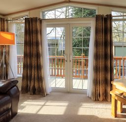 A living room with a leather armchair a floor lamp and a wooden side table with a lamp by glass doors with curtains at Squirrels Snug in Pony Meadow 1 White Cross Bay near Windermere
