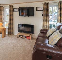 A living room with a brown leather sofa and two pillows a wall mounted television an electric fireplace a wooden side table with a lamp and patterned curtains at Squirrels Snug in Pony Meadow 1 White Cross Bay near Windermere