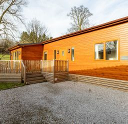 A wooden cabin with a porch and steps surrounded by gravel and trees at Comraich Beckside 40 Limefitt Park near Windermere