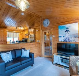 A living room with a black sofa and two cushions near a TV and a kitchen area in a wooden cabin at Lakeshaven Skiptory Howe 16 White Cross Bay near Windermere