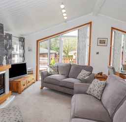 A living room with gray sofas a fireplace a wooden TV stand and large windows at Limefitt Lodge Fellside 8 Limefitt Park near Windermere