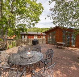 An outdoor wooden deck with metal tables and chairs near wooden lodges and trees at Waterbird Lakeview 4 White Cross Bay near Windermere