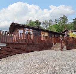 A wooden cabin with a fenced porch and stairs on a gravel area surrounded by trees at Pheasant's Nest Kirkstone 11 Limefitt Park near Windermere