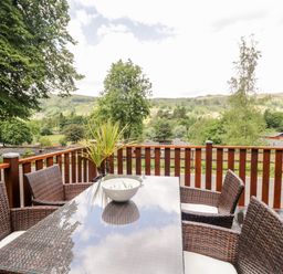 An outdoor wooden deck with a glass top table and wicker chairs overlooking trees and hills at Ambleside Lodge Fellside 3 Limefitt Park near Windermere