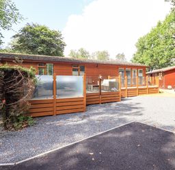 A wooden lodge with a fenced patio area and outdoor furniture surrounded by trees at Lakes Lodge Keswick Court 3 White Cross Bay near Windermere