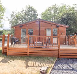 A wooden cabin with a deck and outdoor chairs surrounded by trees at Lakes Lodge Keswick Court 3 White Cross Bay near Windermere