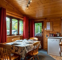 A wooden dining room with a table set for four next to windows with red curtains and a door near kitchen appliances at Glaramara Number 7 Burnside Park near Keswick