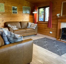 A living room with brown leather sofas and plaid cushions wood panel walls a window with red curtains a fireplace with flowers and a black stove at Glaramara Number 7 Burnside Park near Keswick
