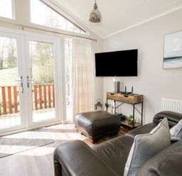 A living room with leather sofa and ottoman by glass doors and TV on wall at Troutbeck Lodge in Beckside 6 Limefitt Park near Windermere