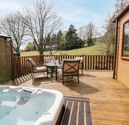 An outdoor wooden deck with a hot tub and a table with four chairs and a rural landscape at Troutbeck Lodge Beckside 6 Limefitt Park near Windermere