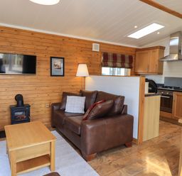 A living room with a brown leather sofa a wooden coffee table a television on a wood paneled wall and a kitchen area at Ramblers Rest Lodge Pinewood 22 Limefitt Park near Windermere