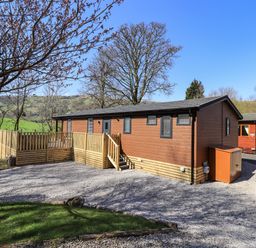 A wooden cabin with a fenced porch and gravel driveway surrounded by trees at Jinnyspinner Lodge Beckside 37 Limefitt Park near Windermere