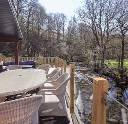 An outdoor dining area with wicker chairs and a table on a deck overlooking a river at Jinnyspinner Lodge Beckside 37 Limefitt Park near Windermere