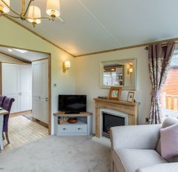 A living room with a sofa, television on a corner unit, fireplace with clock and picture frame on mantel, dining table with purple chairs, and window with curtains at Latterbarrow Lodge in Beckside 46 Limefitt Park near Windermere