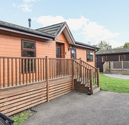 An exterior view of two wooden lodges with decks and steps on a paved area with grass at Sunny Brow Lodge Lakeside 5 White Cross Bay near Windermere