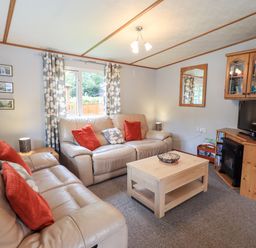 A living room with two beige sofas with red and patterned cushions a wooden coffee table a TV on a wooden cabinet and a window with patterned curtains at Pinehaven Lodge Grasmere 6 White Cross Bay near Windermere