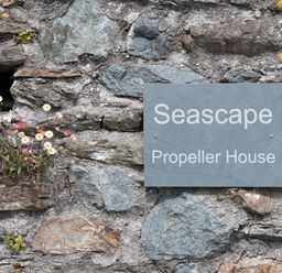 A stone wall with a sign reading Seascape Propeller House and flowers growing in the crevices