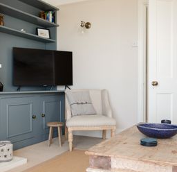 A living room with a TV on blue shelves a beige armchair a wooden coffee table and decorative items at Seascape in Salcombe