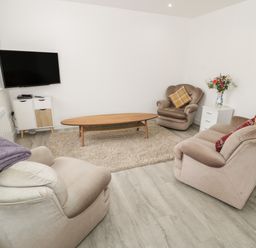 A living room with beige armchairs a wooden coffee table a wall mounted television and a small white cabinet with flowers on a side table at Trigfa in Llanfechell