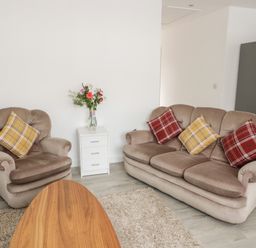 A living room with a beige armchair and sofa with plaid cushions a white drawer unit with flowers and a wooden coffee table at Trigfa in Llanfechell