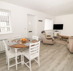 A dining area with a round wooden table set with placemats and glasses next to a living room with two armchairs a wall-mounted TV and a coffee table at Trigfa in Llanfechell