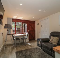 A living room with a dark sofa with animal pillows a small dining table with two chairs a floor lamp a wall-mounted television and a window with a wooden door at The Retreat in Washford near Williton