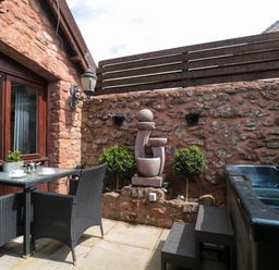 An outdoor patio with a glass table and wicker chairs a stone wall with plants a modern water feature and a hot tub at The Retreat in Washford near Williton
