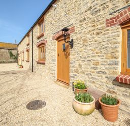 Exterior of a stone house with wooden doors and windows and flower pots on gravel at The Stables Burton Bradstock