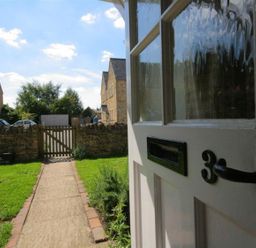 A pathway leading from a partially open door to a wooden gate and stone wall with houses and cars in the background at Noel Cottage in Chipping Campden